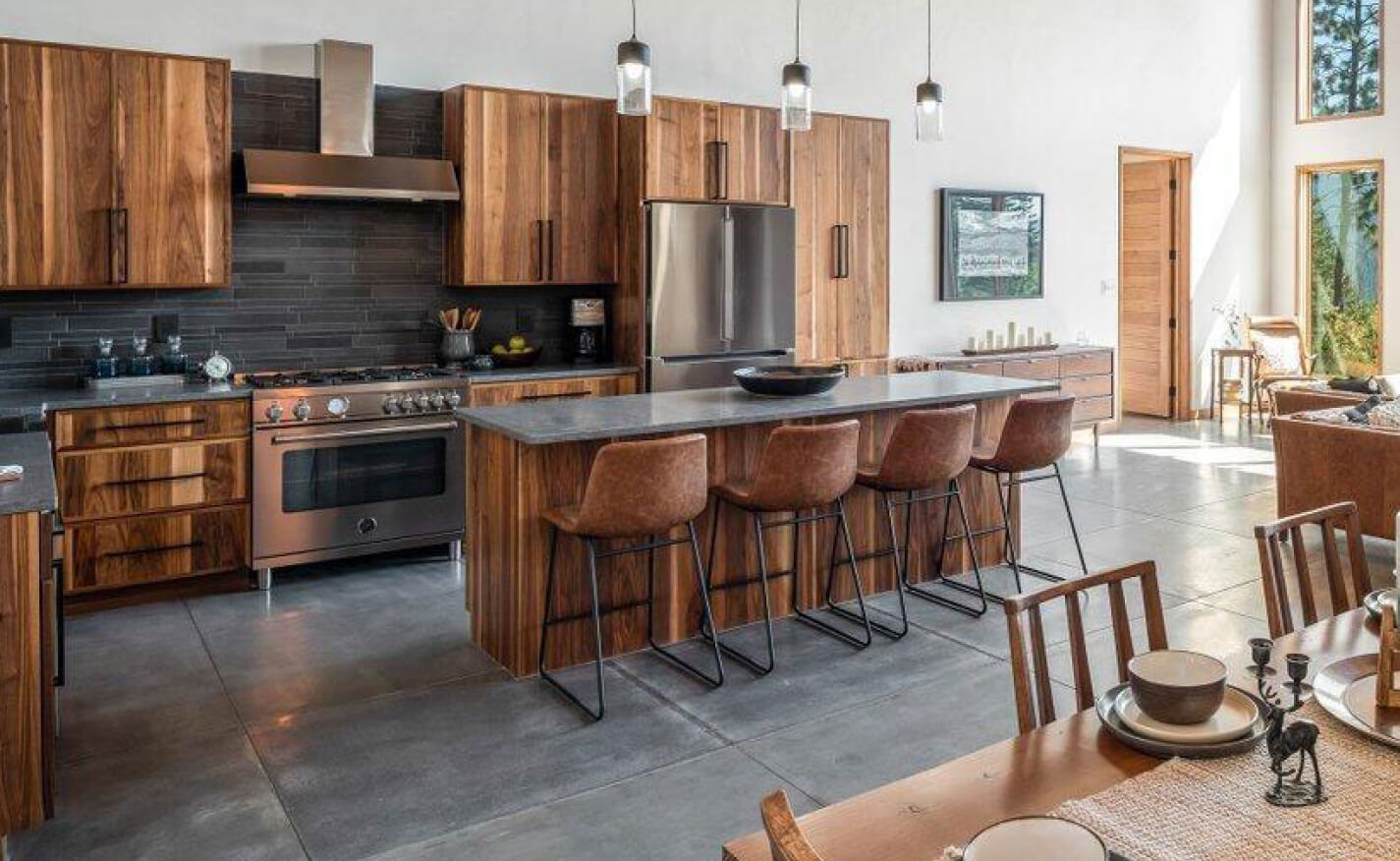 Modern kitchen with wood cabinets, stainless steel appliances, a large island with four brown barstools, and pendant lights. A stacked stone accent enhances the space, while large windows fill the room with natural light.