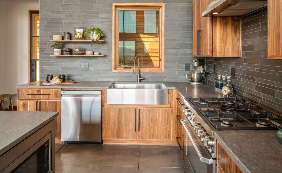 Modern kitchen with wooden cabinets, stainless steel appliances, farmhouse sink, gas stove, gray tile backsplash, high end stone design accents, open shelves with dishes and plants, and a window above the sink letting in natural light.
