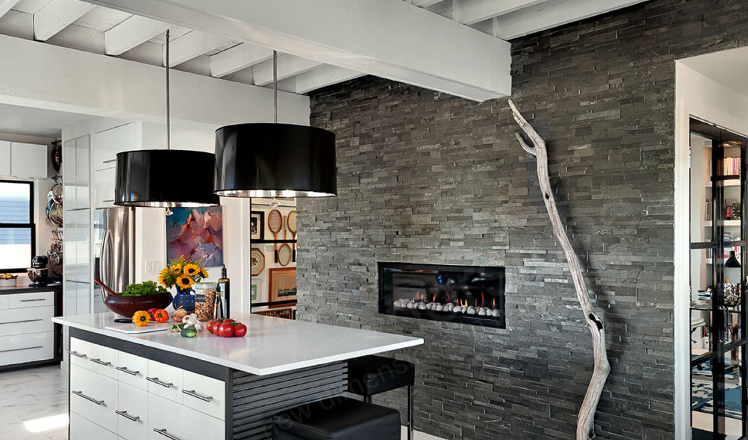 Modern kitchen with a white island, black pendant lights, and a stacked stone accent wall featuring a sleek fireplace and a tall, decorative branch; sunflowers and fresh produce adorn the island.