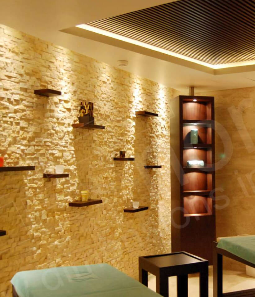 A modern spa room with a stacked stone accent wall, floating shelves displaying decorative items, soft lighting, and two padded massage tables in the foreground.