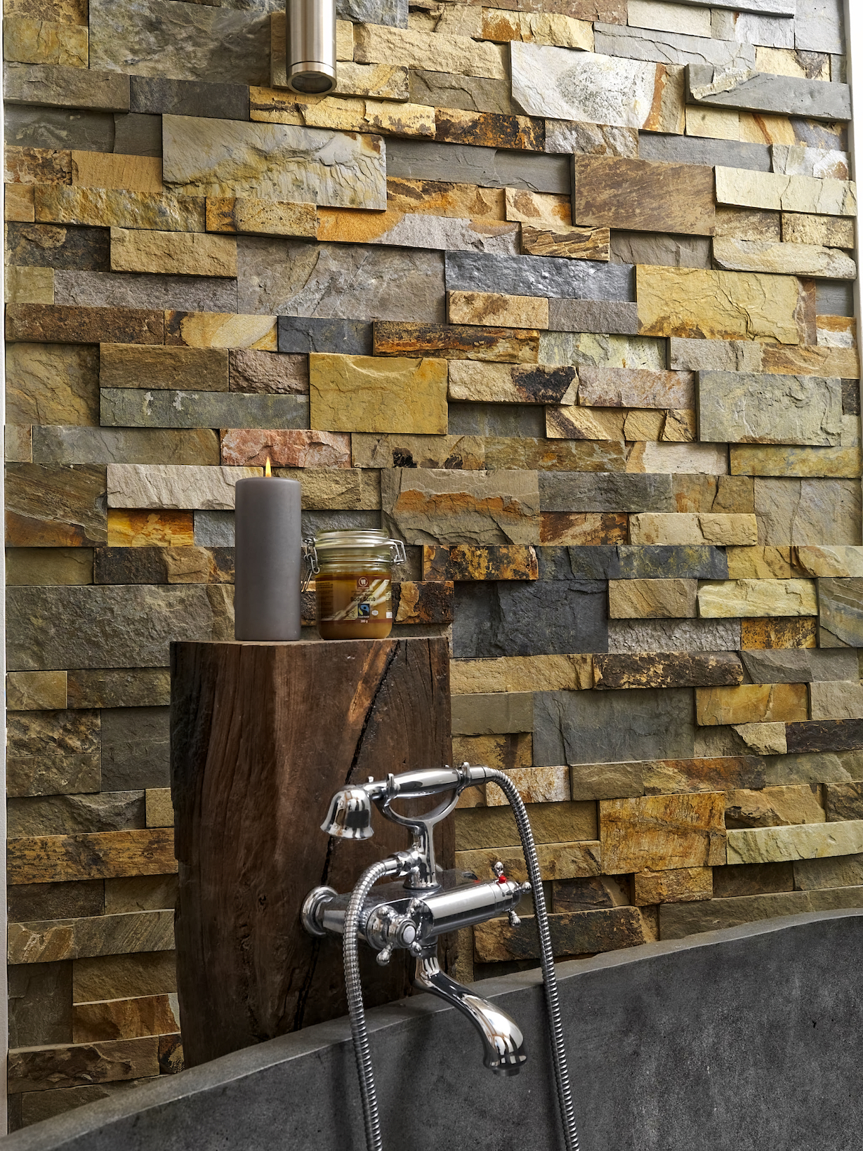 A rustic bathroom features a stone veneer wall, a vintage-style silver faucet with attached shower hose, and a wooden stand holding a gray candle and a glass jar.
