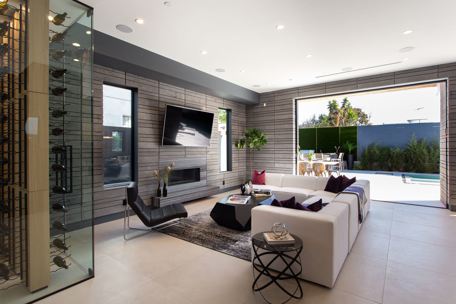 Modern living room with a white sectional sofa, black accent chair, wall-mounted TV, and glass wine storage. A stone accent wall adds elegance. Large sliding doors open to a sunny patio with outdoor seating and a pool.