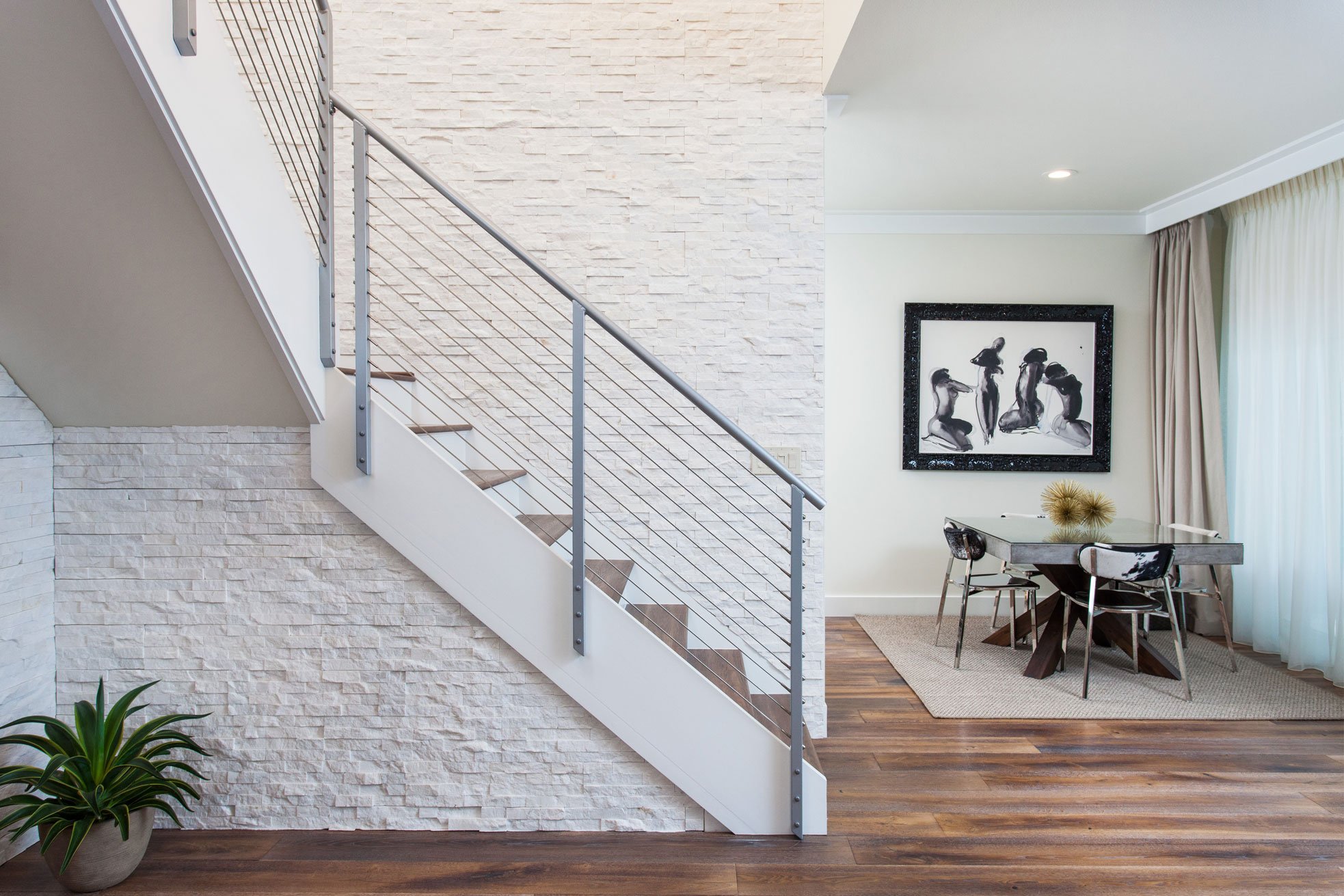 Modern interior with a stacked stone accent wall and staircase with metal railing, wooden floor, a small dining table for four, a plant in the corner, large window with curtains, and black-and-white artwork on the wall.