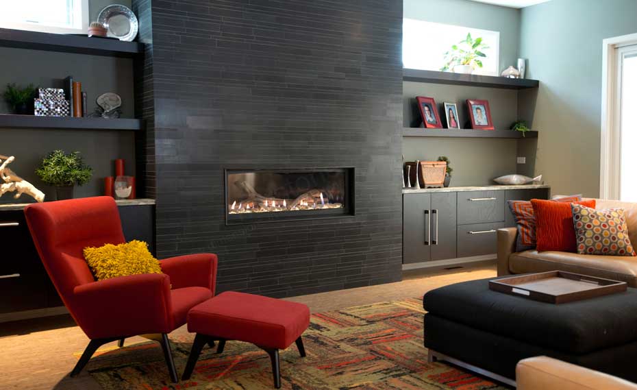 A modern living room featuring a sleek black tile fireplace, red armchair with matching footrest, patterned rug, brown sectional sofa, and shelves displaying decorative items. A natural stone veneer accent wall adds high-end stone design to the space.