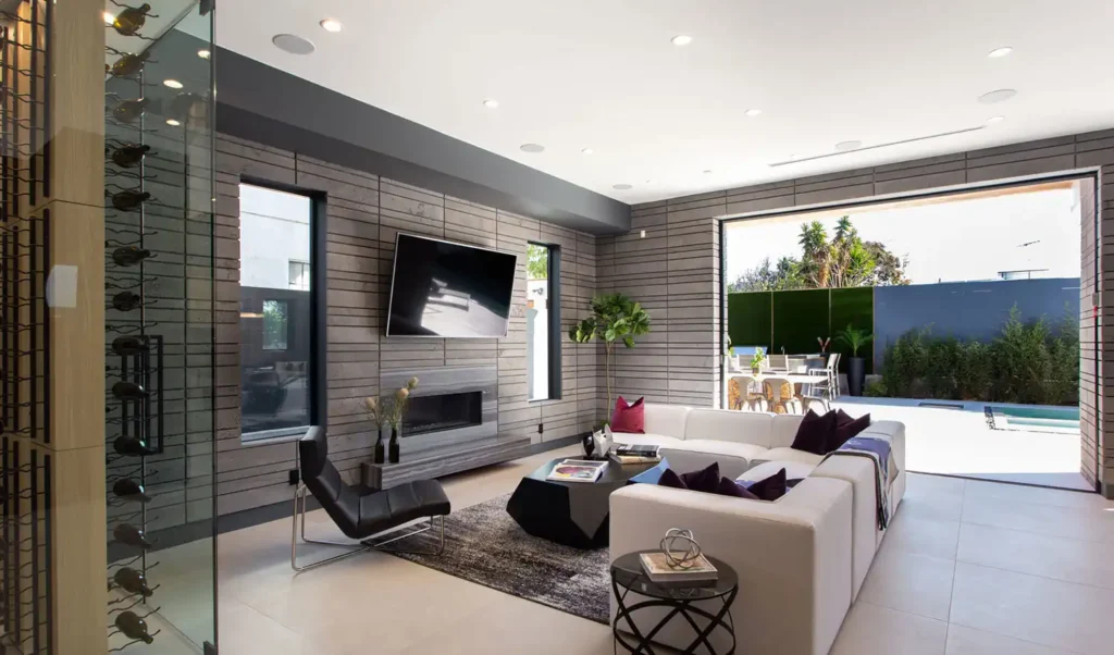 Modern living room with a stone accent wall, mounted TV above a fireplace, a white sectional sofa with dark cushions, black lounge chair, and large sliding doors opening to a sunny outdoor patio and garden.
