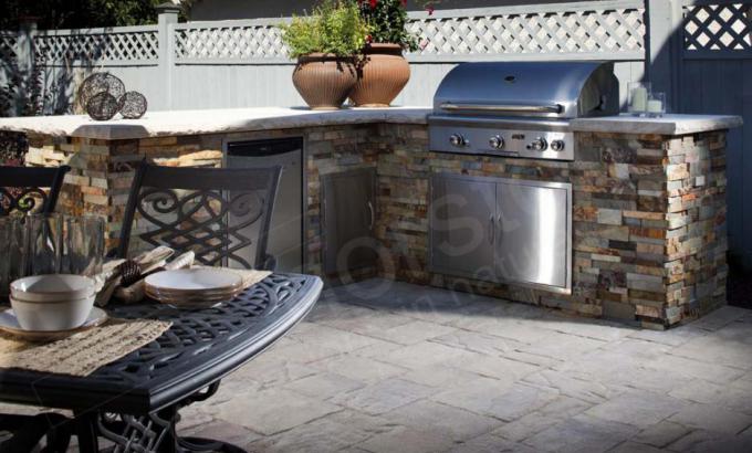 Outdoor kitchen with a high end stone design counter, built-in grill, mini fridge, and plants in large pots. In the foreground, a metal patio table is set with dishes on a tiled patio, and a white fence surrounds the area.