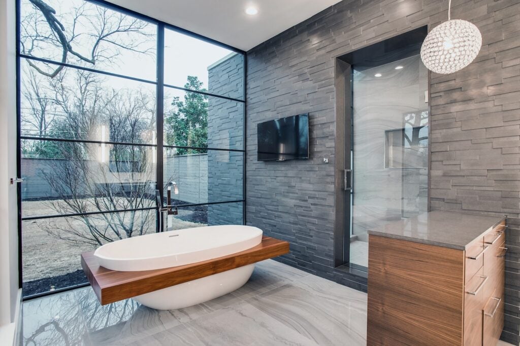 Modern bathroom with a freestanding white tub featuring a wooden accent, large floor-to-ceiling windows, gray stacked stone wall tiles, a mounted TV, wooden cabinets, and a spherical pendant light.