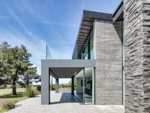 Modern two-story house with large glass windows, high end stone design and brick exterior, flat roof, and spacious patio, set on a sunny day with green grass and trees in the background.