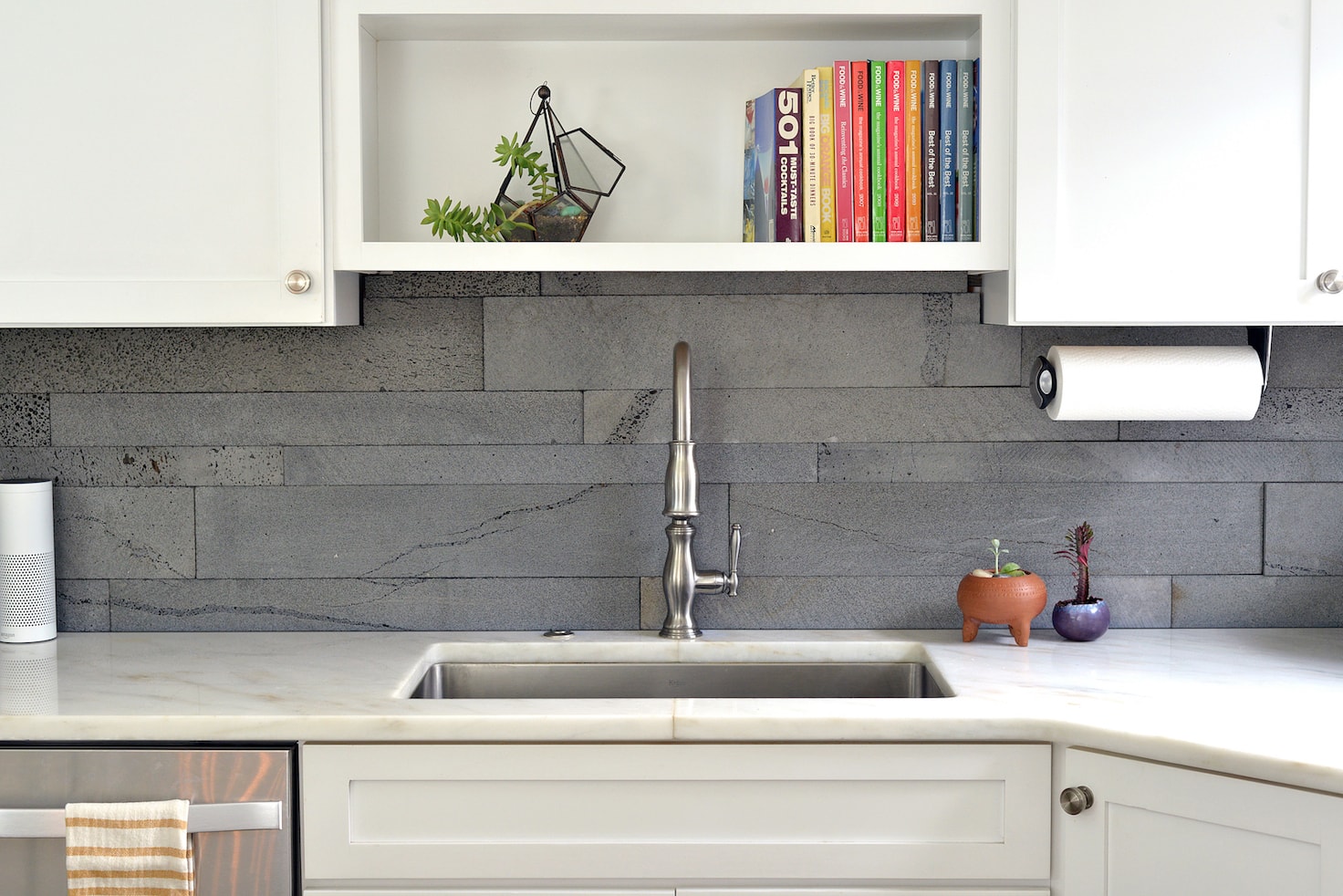 Modern kitchen sink area with natural stone veneer backsplash, silver faucet, white cabinets, and open shelf holding books and small potted plants. A paper towel holder is mounted under the shelf, and countertops are white.