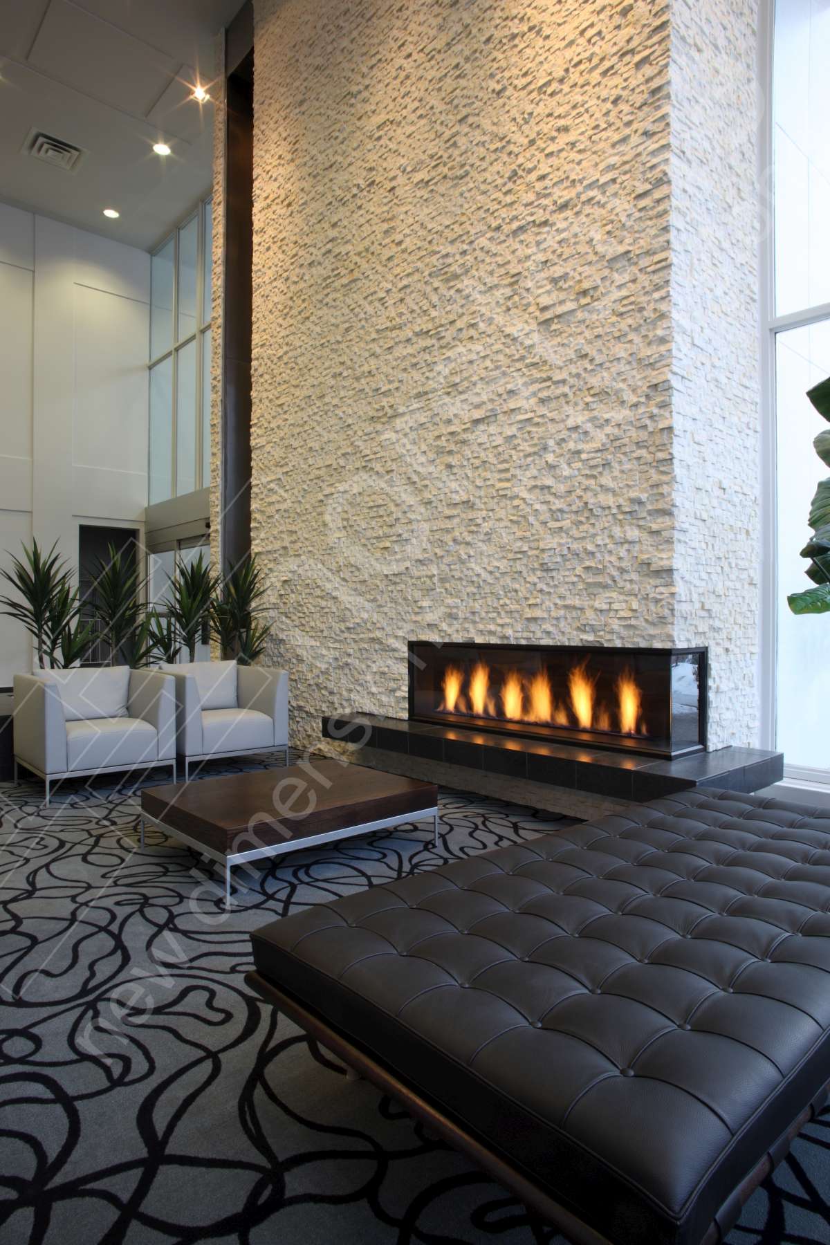 Modern lounge area with white chairs and black ottoman, a patterned carpet, large natural stone veneer accent wall, and a sleek linear fireplace beneath tall windows; potted plants add greenery.