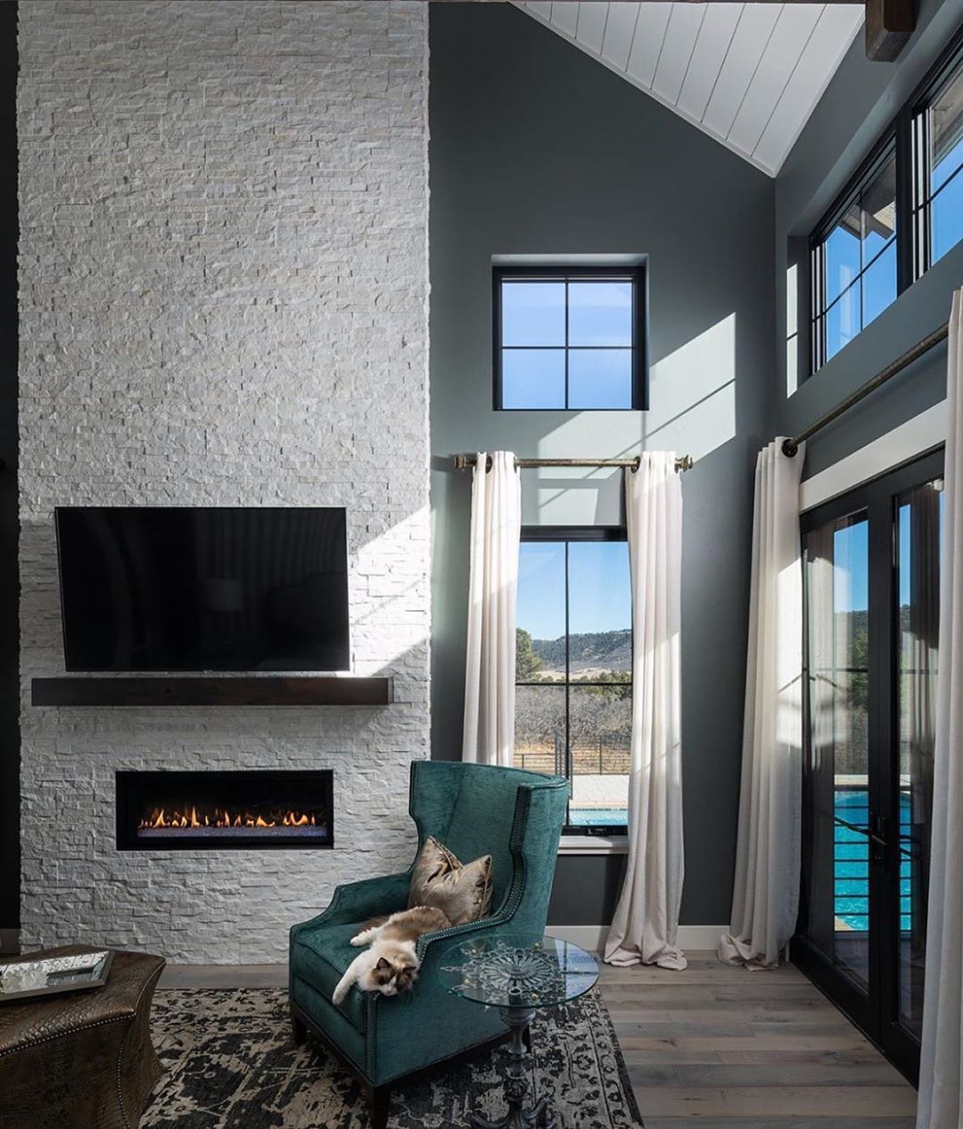 A cozy modern living room with high ceilings, large windows, white curtains, a stacked stone fireplace with a TV above, a teal armchair where a cat is sleeping, and sunlight streaming in.