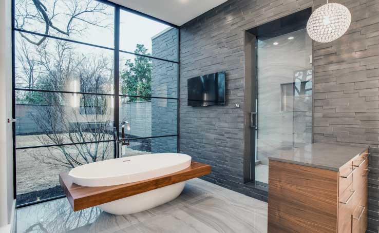 Modern bathroom with a large oval bathtub, floor-to-ceiling windows, gray natural stone veneer walls, glass shower, wall-mounted TV, wooden vanity, and a spherical pendant light. Trees are visible outside.