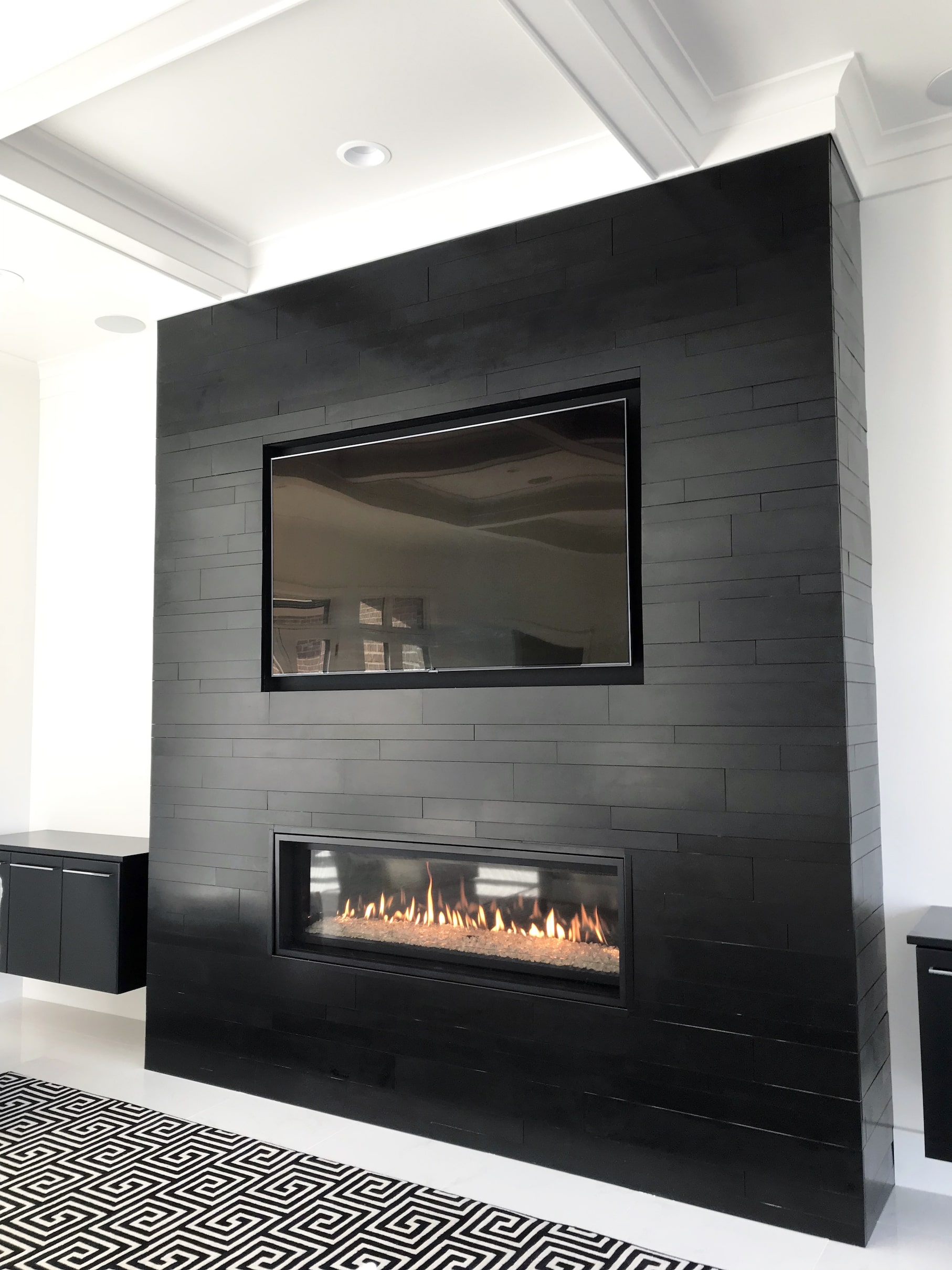 A modern black tiled wall features a built-in flat-screen TV above a rectangular gas fireplace with visible flames and a natural stone veneer surround, set in a bright room with white walls and a geometric black-and-white rug.
