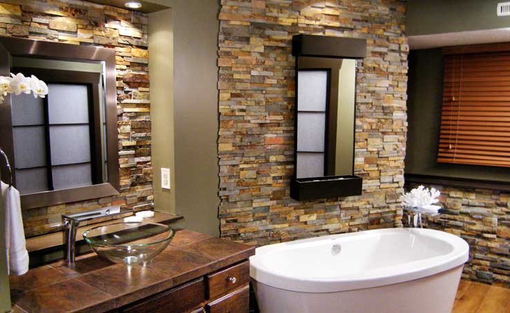 A modern bathroom with a freestanding white bathtub, glass vessel sink on a wooden vanity, natural stone veneer accent walls, two mirrors, and wooden blinds on a window.