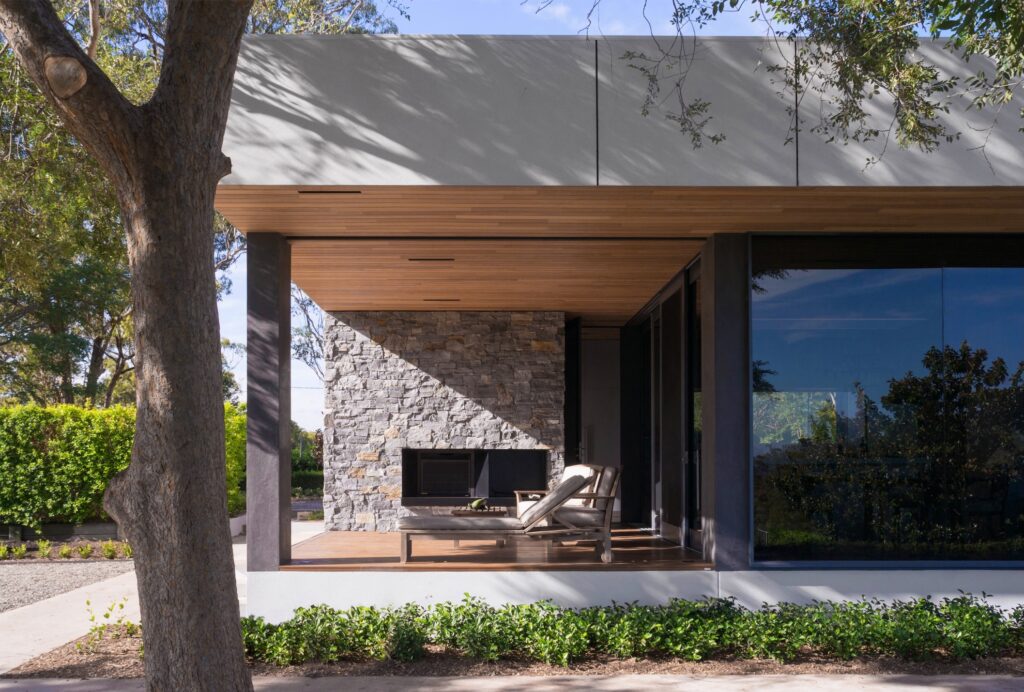 Modern house exterior with large windows, a wooden patio ceiling, and a lounge chair facing a stunning stone outdoor fireplace—perfect inspiration for unique stone outdoor fireplace ideas—surrounded by lush greenery and trees under a clear sky.