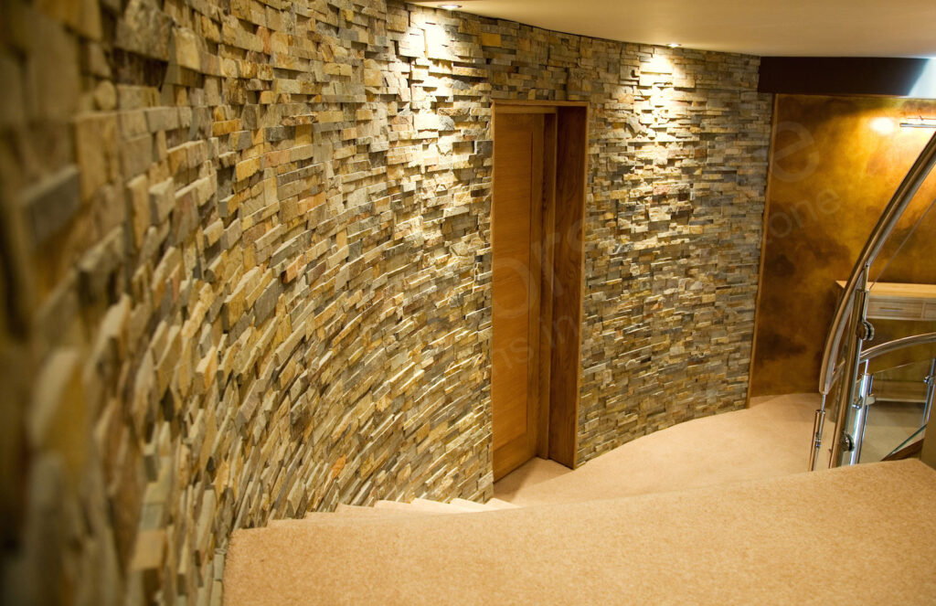 Curved staircase with beige carpeting, stone accent wall, wooden door, and warm spotlights creating a cozy, modern atmosphere. Glass and metal railing is partially visible on the right.
