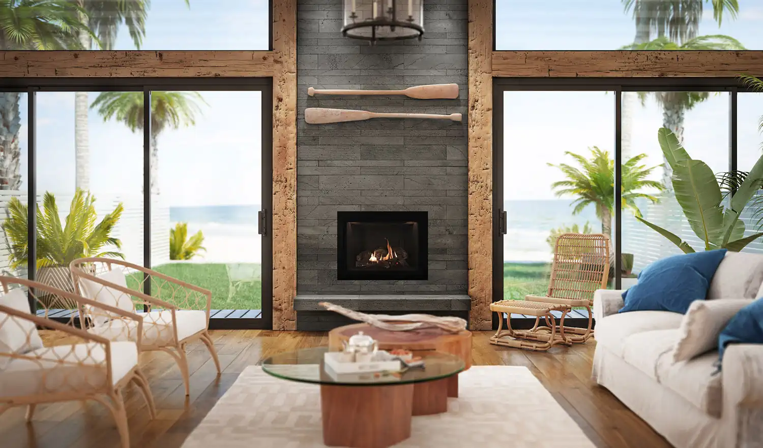 A bright coastal living room with large windows, wicker furniture, tropical plants, and a wooden coffee table features a stone fireplace set in a gray wood accent wall with two decorative oars above it. Ocean and palm trees are visible outside.